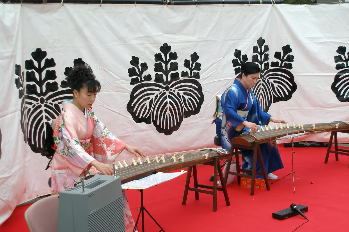 Two women in kimono play koto on trestle stands at a public performance, with a paulownia crest on the white banner behind them