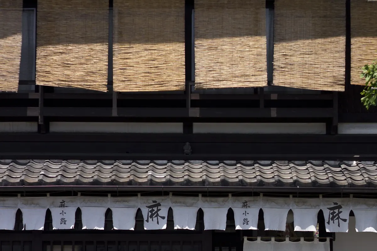 The front of a Kyoto machiya townhouse, with woven bamboo screens on the upper floor, a tiled eave and white noren curtains above a lattice window
